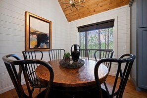 Un coin repas avec une table et des chaises en bois, une œuvre d’art encadrée au mur et une fenêtre donnant sur des arbres.