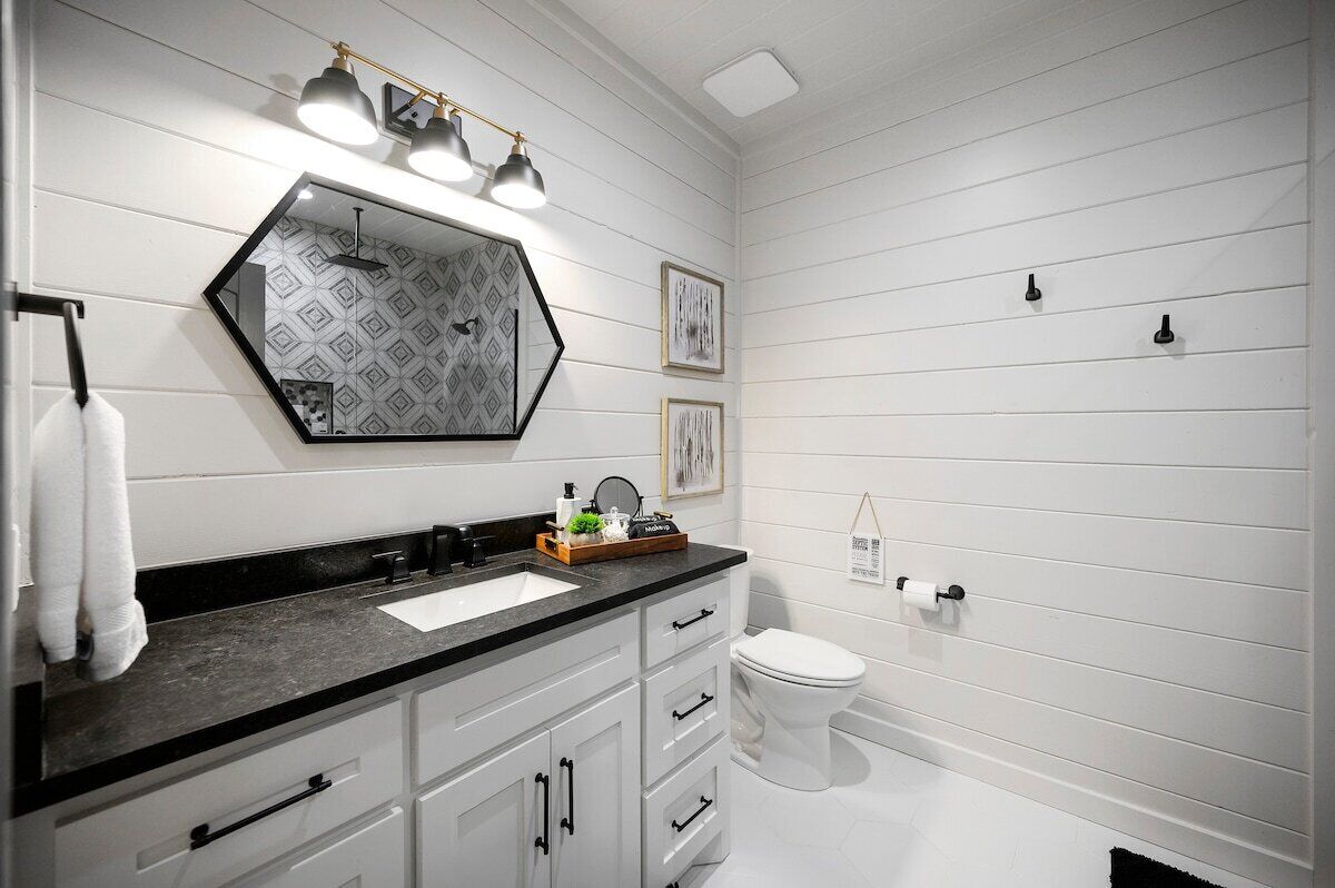 Une salle de bain avec un plan de toilette noir, des meubles blancs, des toilettes blanches et un miroir orné d’un motif géométrique.