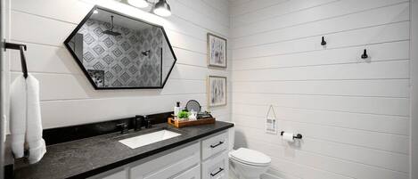 Une salle de bain avec un plan de toilette noir, des meubles blancs, des toilettes blanches et un miroir orné d’un motif géométrique.