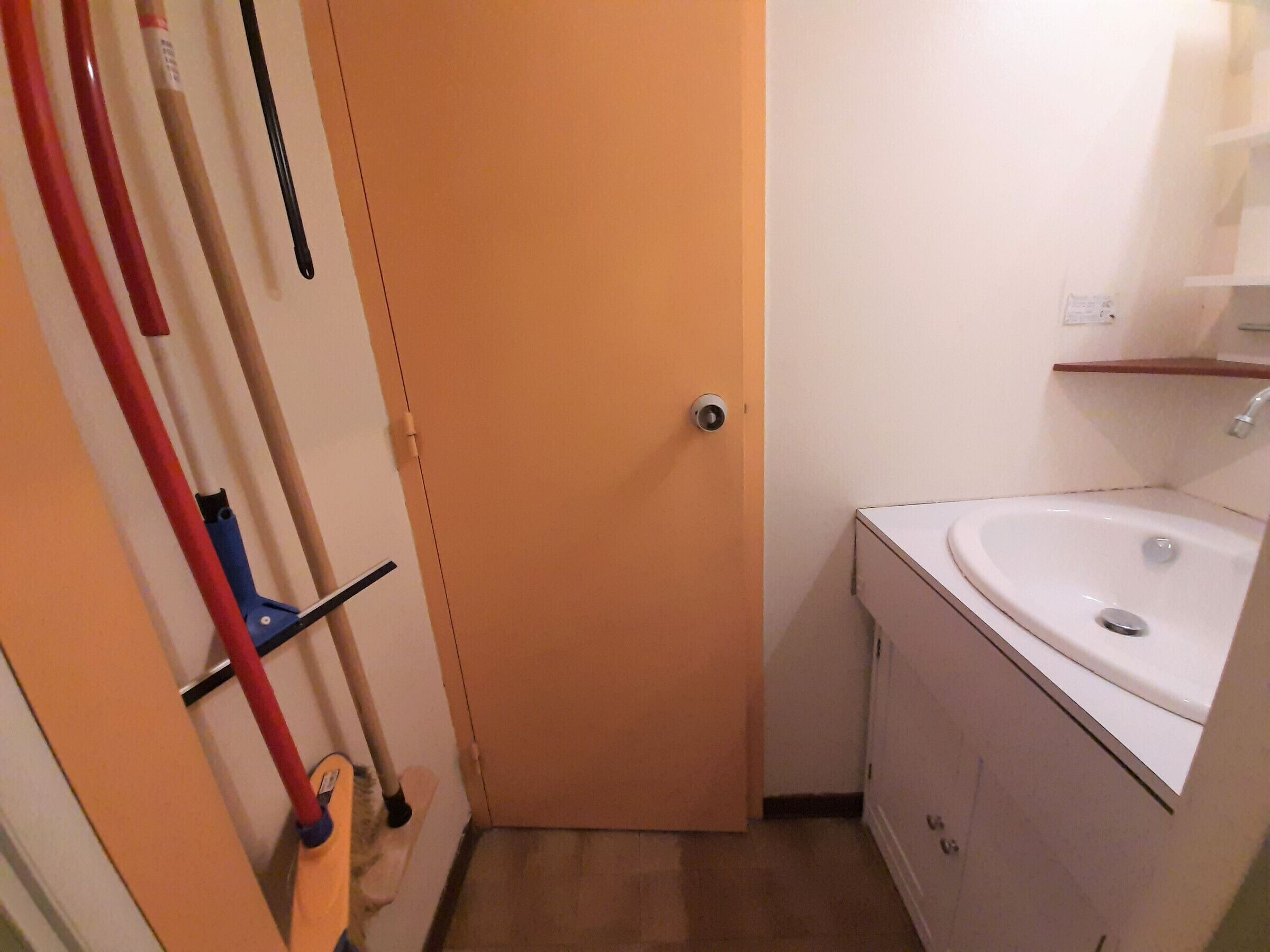 Un baño pequeño con un lavamanos blanco, un espejo y una puerta. Hay herramientas de limpieza colgadas en la pared.