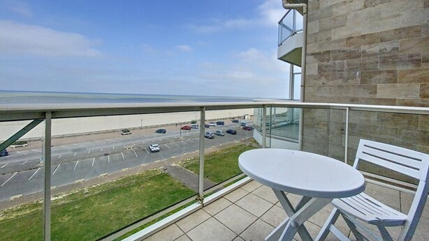 Un balcon avec une table et des chaises, donnant sur un parking et une plage.