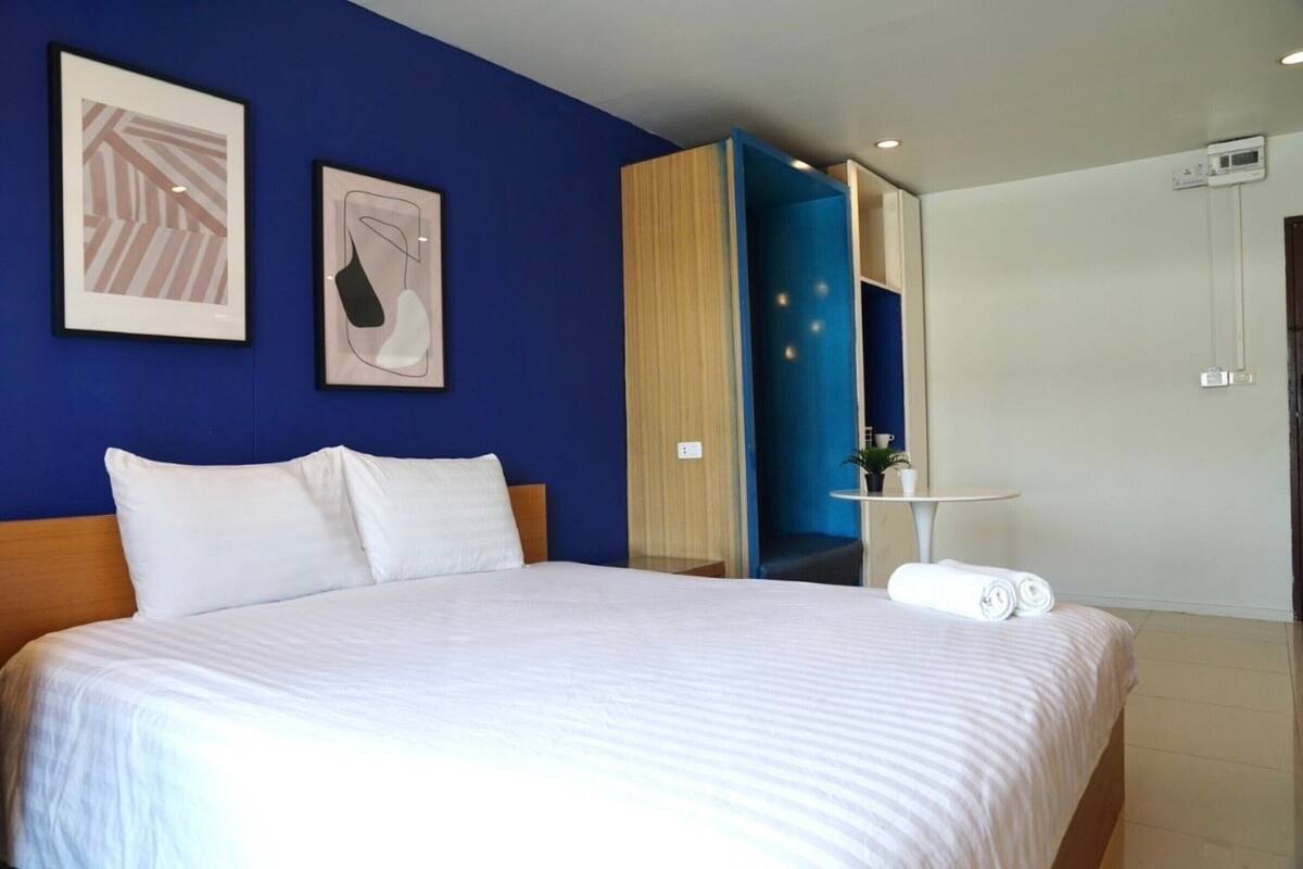 A hotel room with a large bed, two framed artworks on the wall, a small table with a plant, and a bathroom door visible.