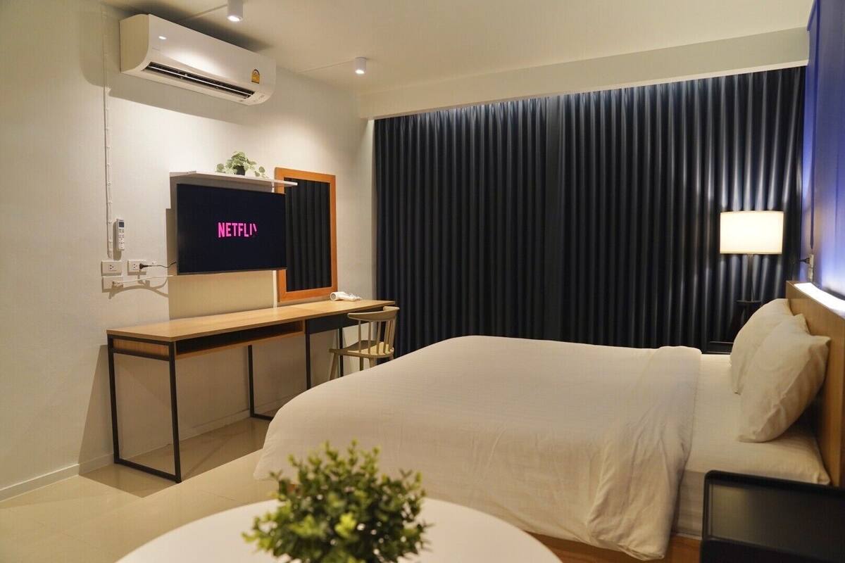 A hotel room with a bed, a desk with a TV displaying 'NETFLIX', a window with dark curtains, and a small plant on the desk.