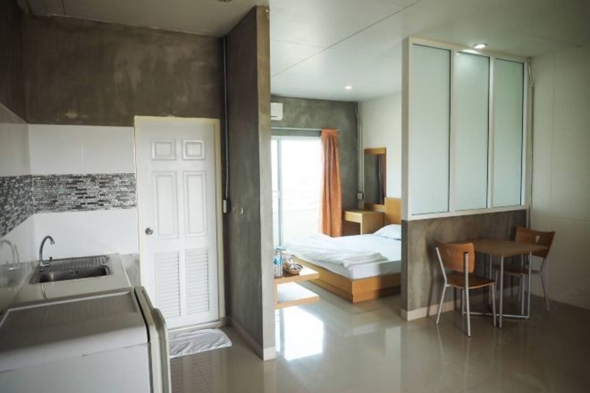 A modern hotel room with a kitchenette, a bed with a wooden headboard, a desk with two chairs, and a large wardrobe.