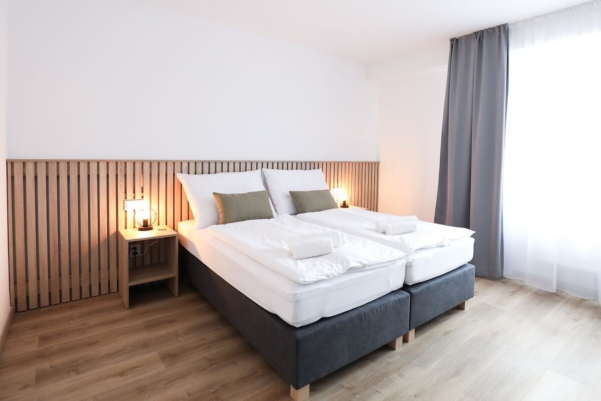 Una habitación de hotel moderna con cabecera de madera, cama con ropa de cama blanca, mesita de noche con lámpara y un ventanal con cortinas.