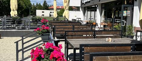 An outdoor seating area with wooden benches and potted plants, a restaurant with outdoor seating, and string lights against a clear sky.