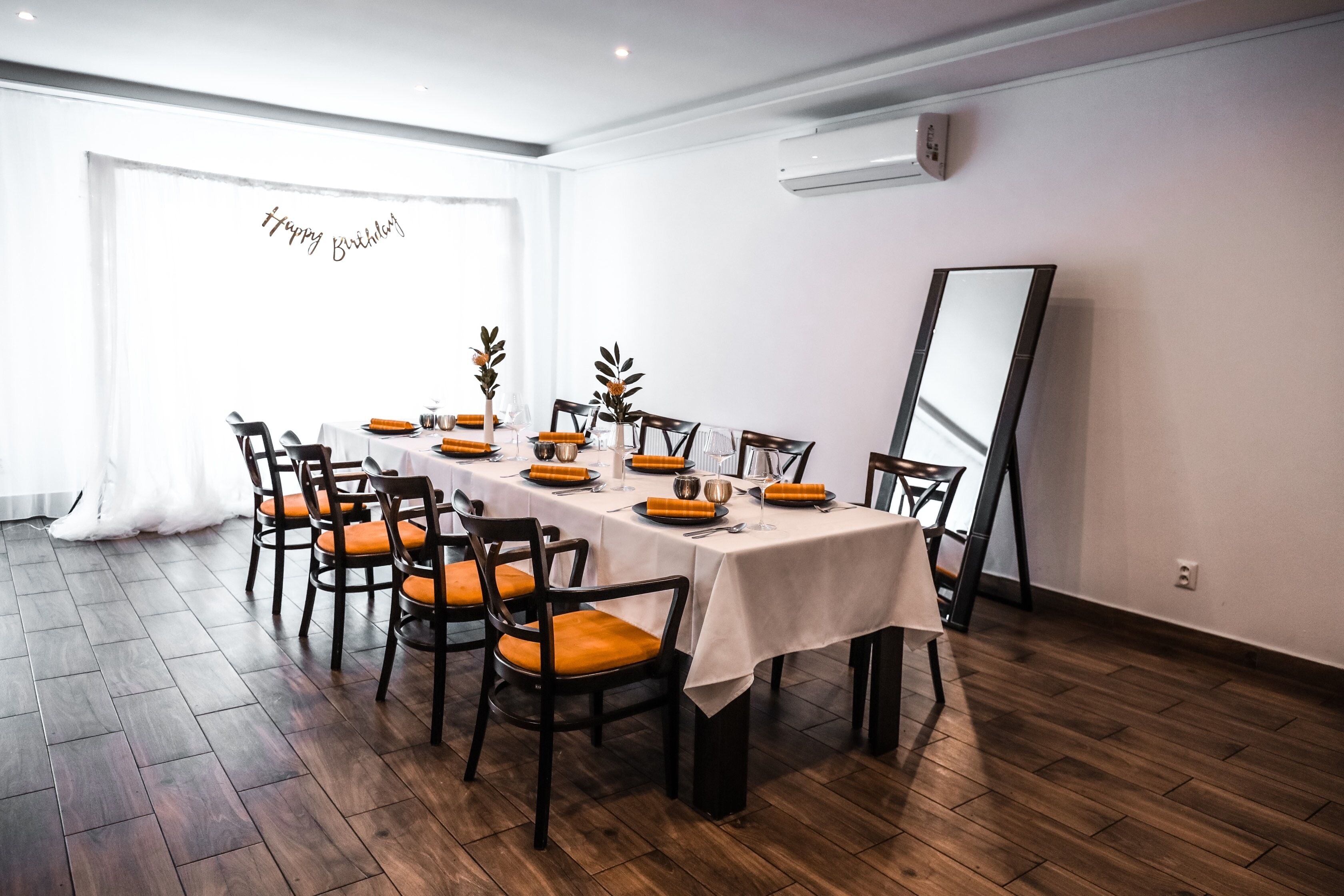 A dining area with a long table set for a meal, surrounded by chairs with orange cushions.