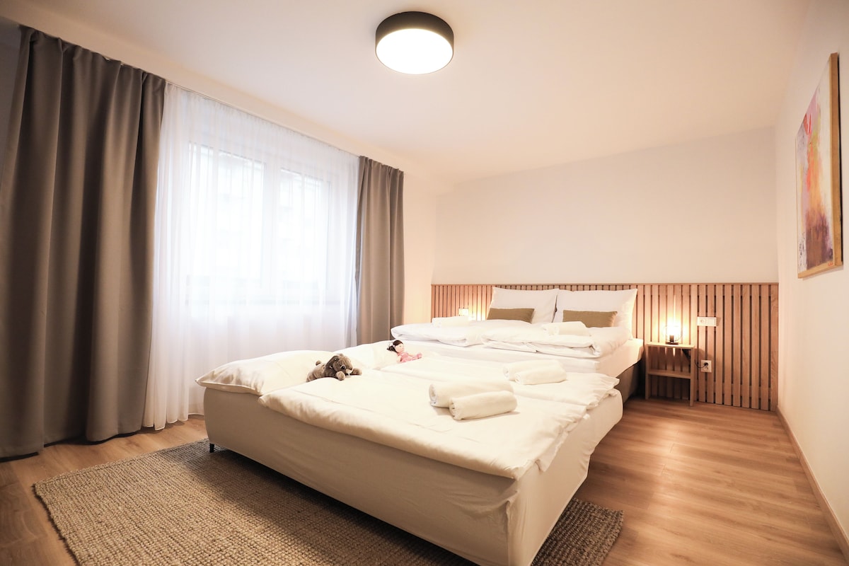 A hotel room with a large bed, a bedside table with a lamp, a wooden headboard, and a large window with curtains.