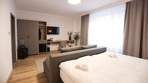 A hotel room with a bed, a sofa, a desk with a TV, a wardrobe, and a plant.