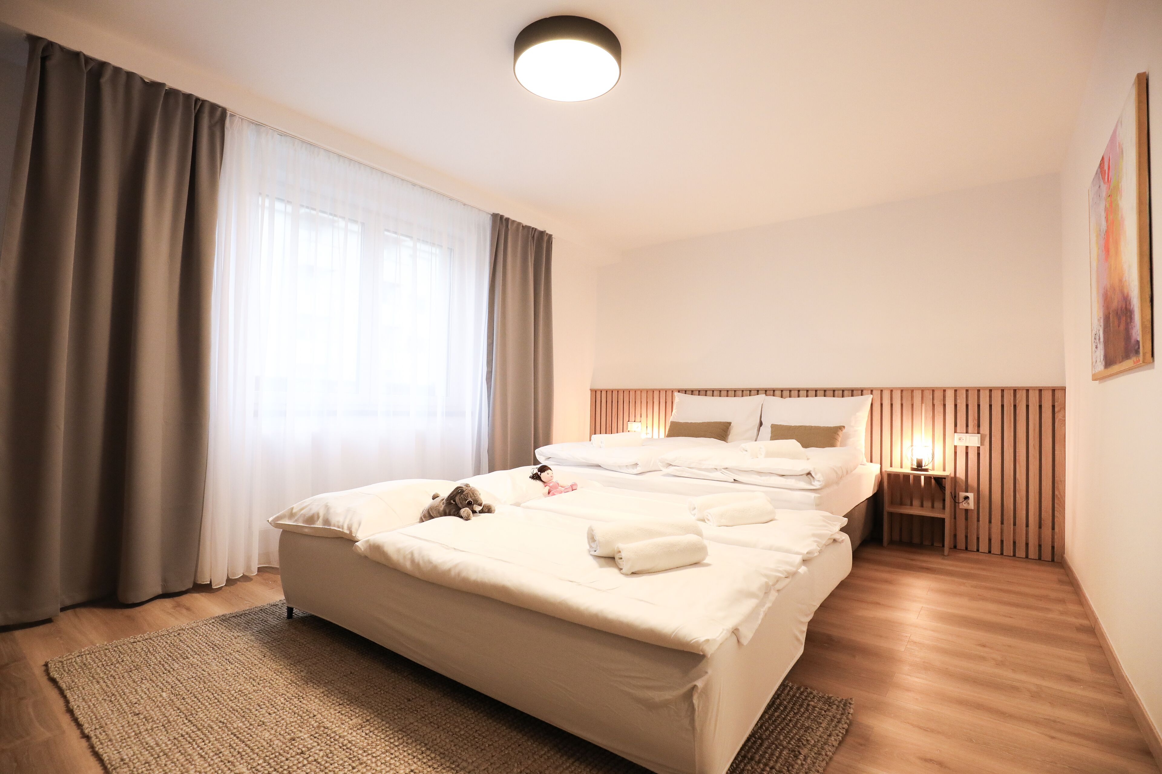Una habitación de hotel con una cama grande, una mesita de noche con lámpara, un cabecero de madera y un ventanal con cortinas.