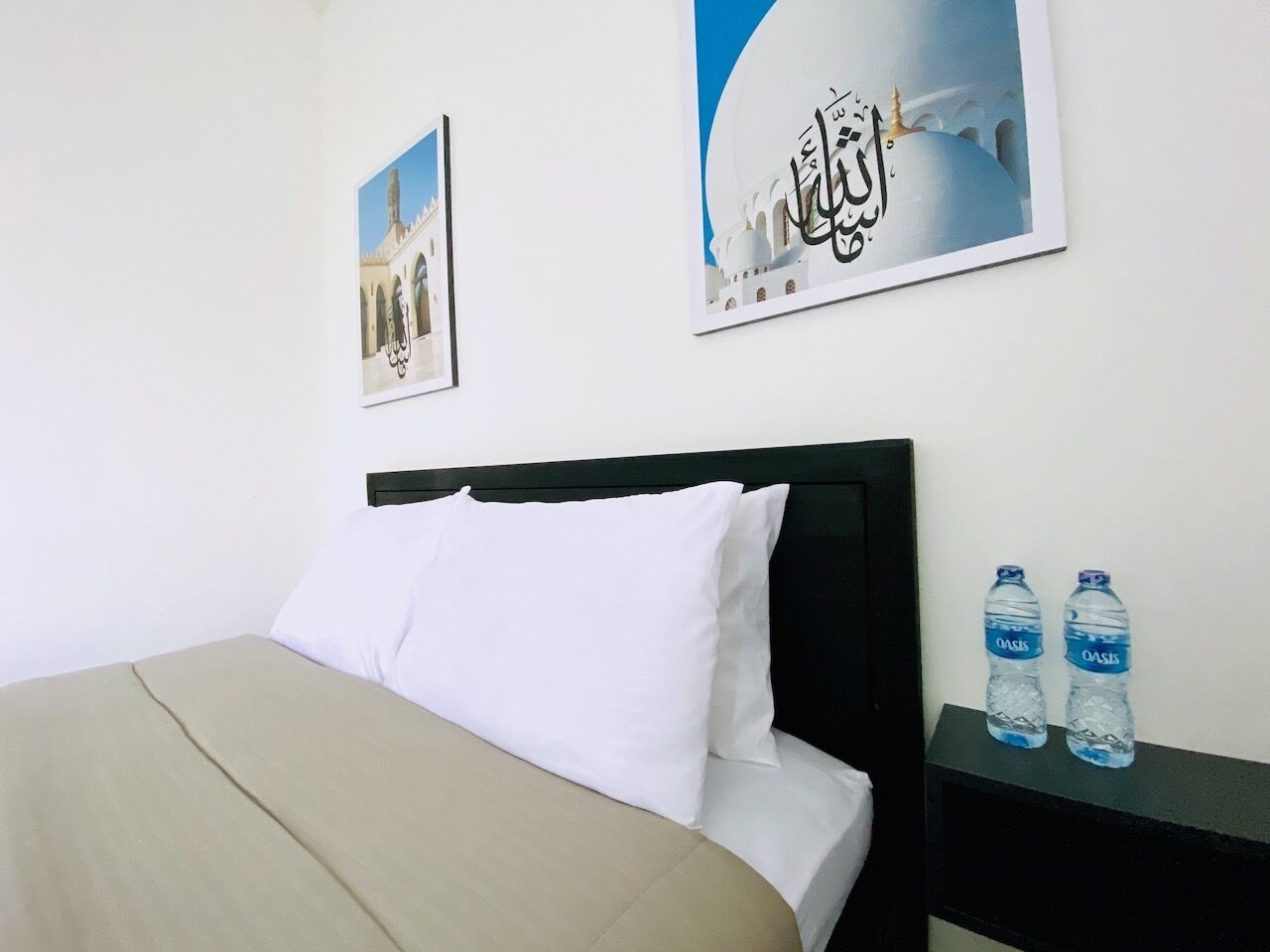 A hotel room with a bed, white pillows, a black headboard, and two water bottles on a nightstand.