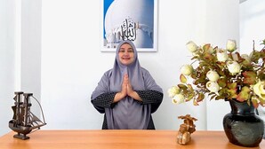 Una persona con un hiyab gris de pie frente a un escritorio con un marco con una imagen de una mezquita, un florero y una pequeña escultura de madera.