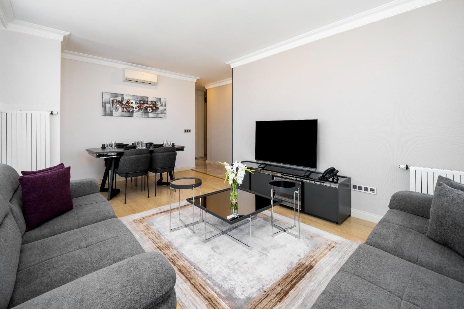 A modern living room with grey sofas, a coffee table, a dining area with chairs, and a flat-screen TV mounted on the wall.