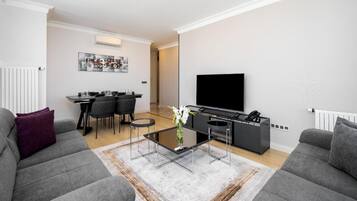 A modern living room with grey sofas, a coffee table, a dining area with chairs, and a flat-screen TV mounted on the wall.