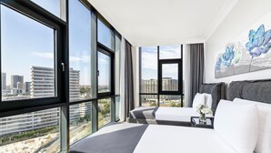 Habitación de hotel moderna con ventanas amplias, dos camas, vista a la ciudad y un cuadro en la pared.