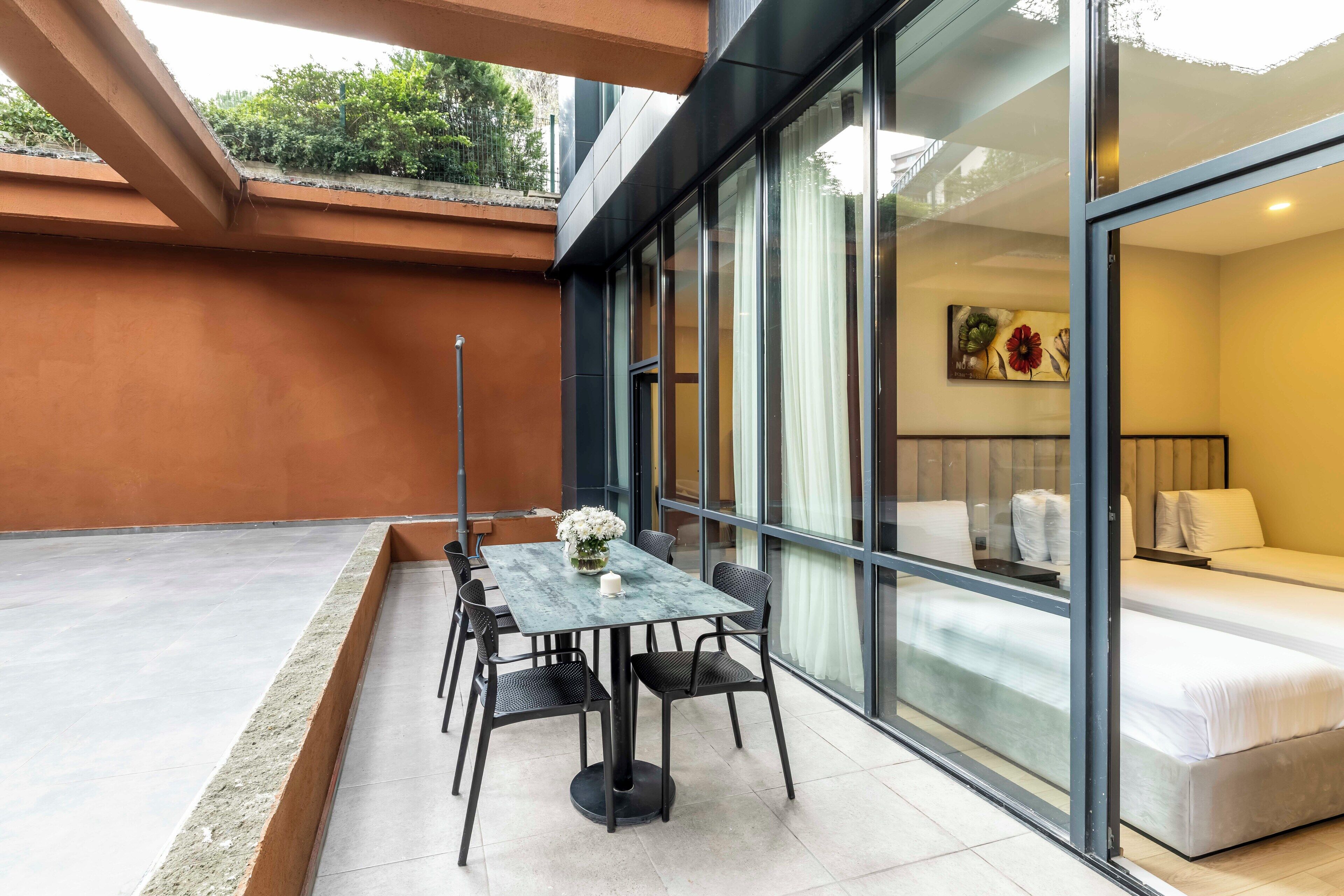 Une chambre d’hôtel moderne avec un mur en verre, un lit, une petite table avec des chaises et un vase à fleurs.
