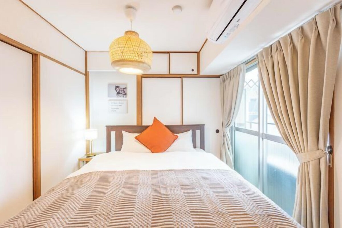 A hotel room with a bed, a bedside table, a lamp, a window with curtains, and a hanging light fixture.