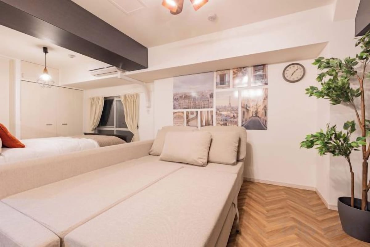 A hotel room with a bed, a window with a city view, a clock on the wall, and a potted plant.