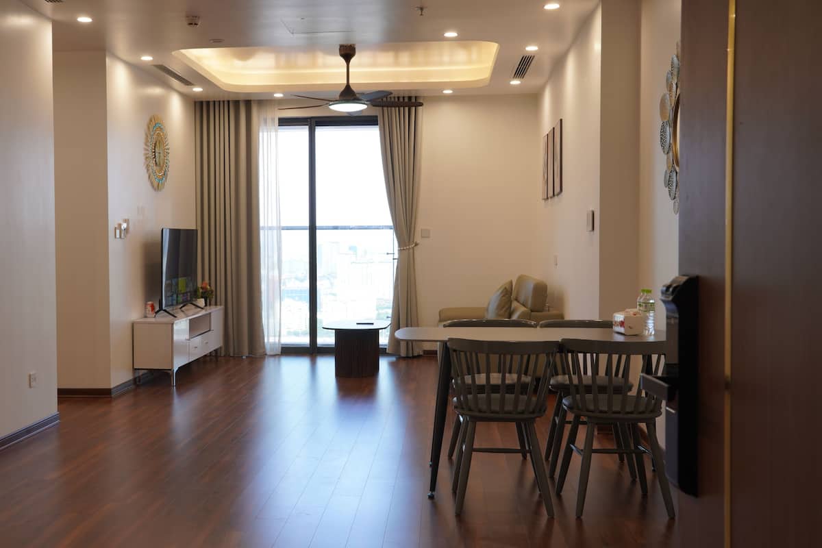 A modern living room with a flat-screen TV, a dining table with chairs, a sofa, and a large window with curtains.