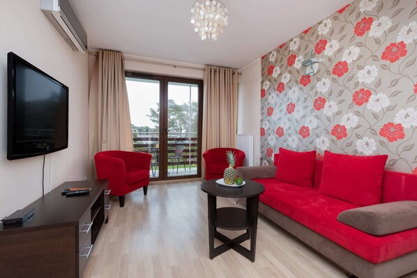Uma sala de estar moderna com televisão de tela plana, sofá vermelho e marrom, uma mesinha com abacaxi e uma varanda com vista.