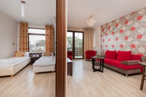 A hotel room with two single beds, a sofa, a dining table, and a balcony with red chairs.
