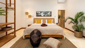 A bedroom with a bed, wooden headboard, two bedside tables, a wall-mounted light, a painting, a door, and a potted plant. - Cozy 4BR Villa With Amazing Pool Area, Bali Villa 2210 (Seminyak)