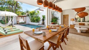 Uma sala de jantar com vista para a piscina, mobiliário de madeira e plantas tropicais.