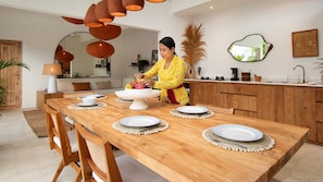 Uma sala de jantar com mesa de madeira posta para quatro pessoas, uma mulher preparando comida e uma cozinha moderna ao fundo.