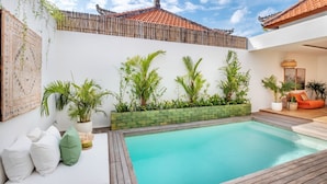 A swimming pool with a wooden deck, surrounded by a white wall with a tiled roof, and various plants. - Elegant 2BR Villa W/ Pool, Bali Villa 2221 (Canggu)