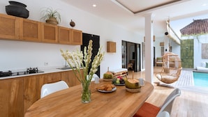 A modern kitchen with wooden cabinets, a dining table with a vase of flowers, and a view of a pool area with a hanging chair. - Elegant 2BR Villa W/ Pool, Bali Villa 2221 (Canggu)