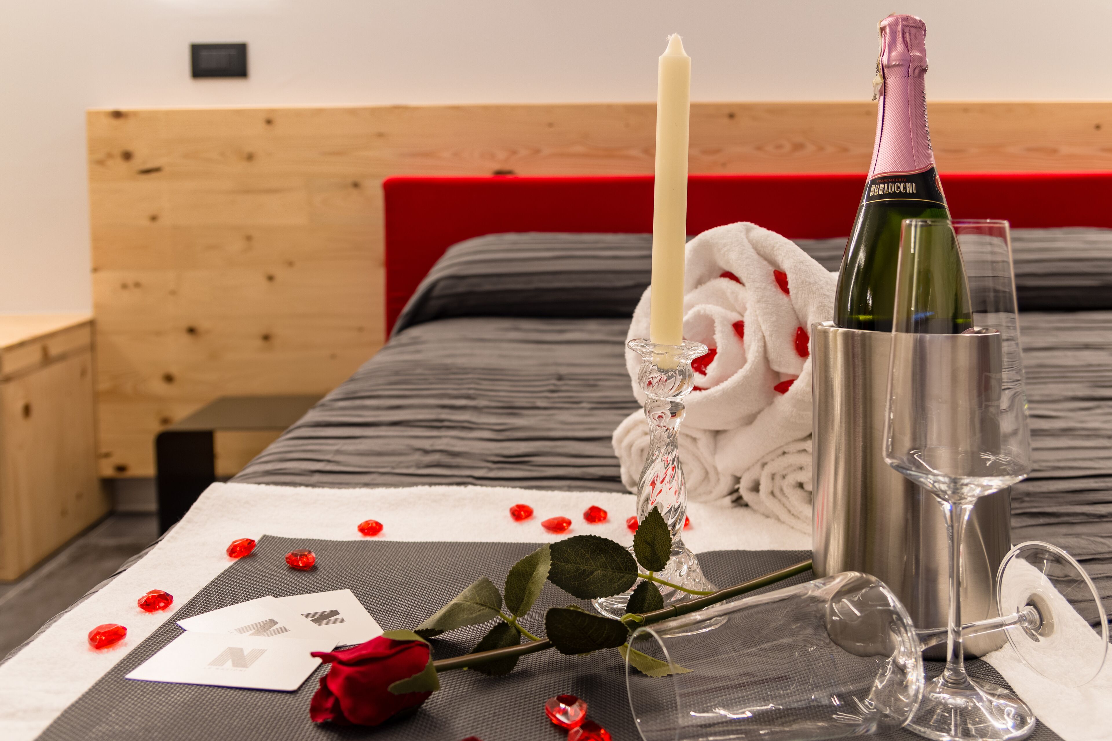 Una cama ordenada con un cabecero rojo, una botella de champán, una copa, una vela, pétalos de rosa y una rosa.