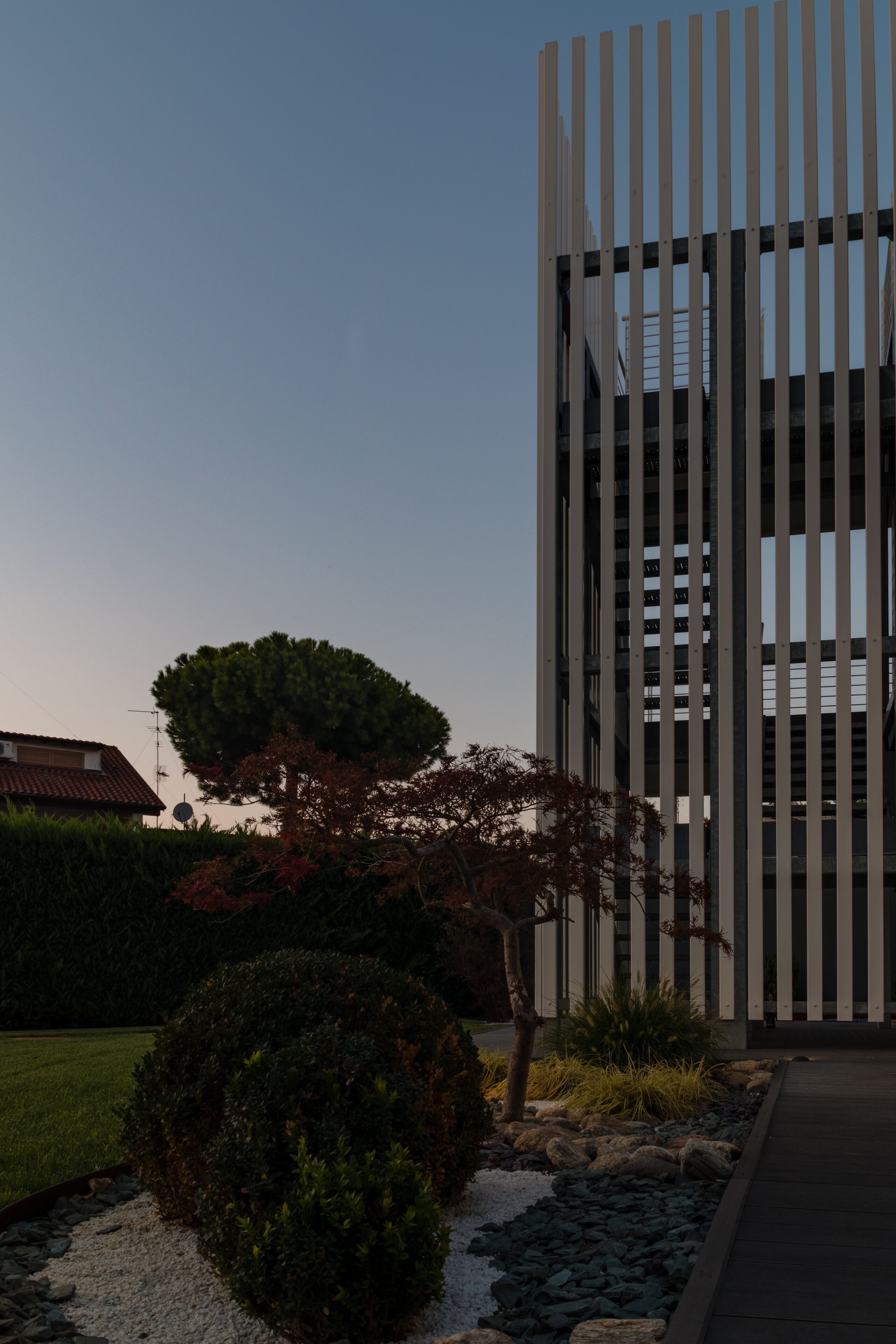 A modern building with vertical lines, surrounded by a landscaped garden with a large tree and a well-maintained lawn.