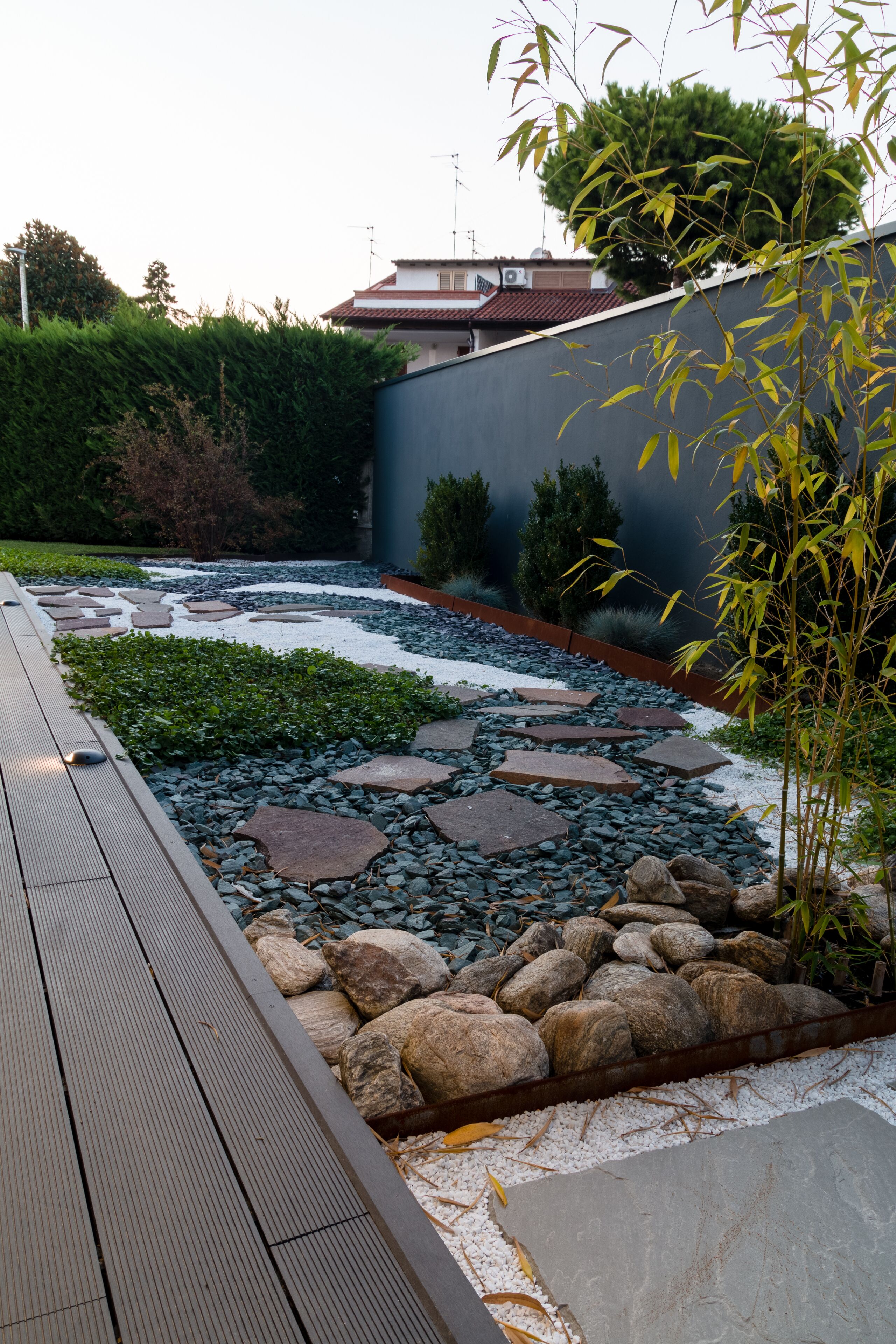 Un jardín con terraza de madera, piedras para pisar, rocas y una planta de bambú.