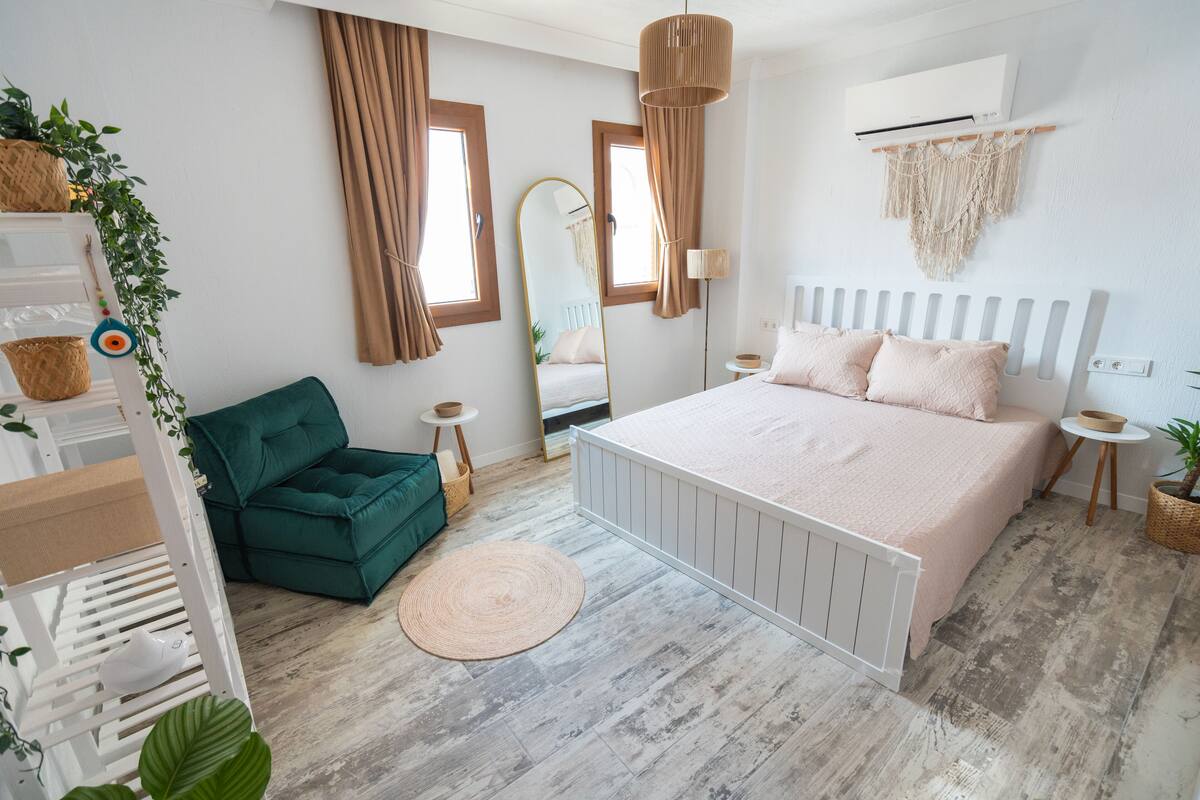Une chambre à coucher avec un lit, un fauteuil vert, un tapis rond, un miroir et une décoration murale en macramé.