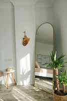 Una habitación con un espejo grande, un taburete pequeño, una planta en maceta y una lámpara fijada a la pared.