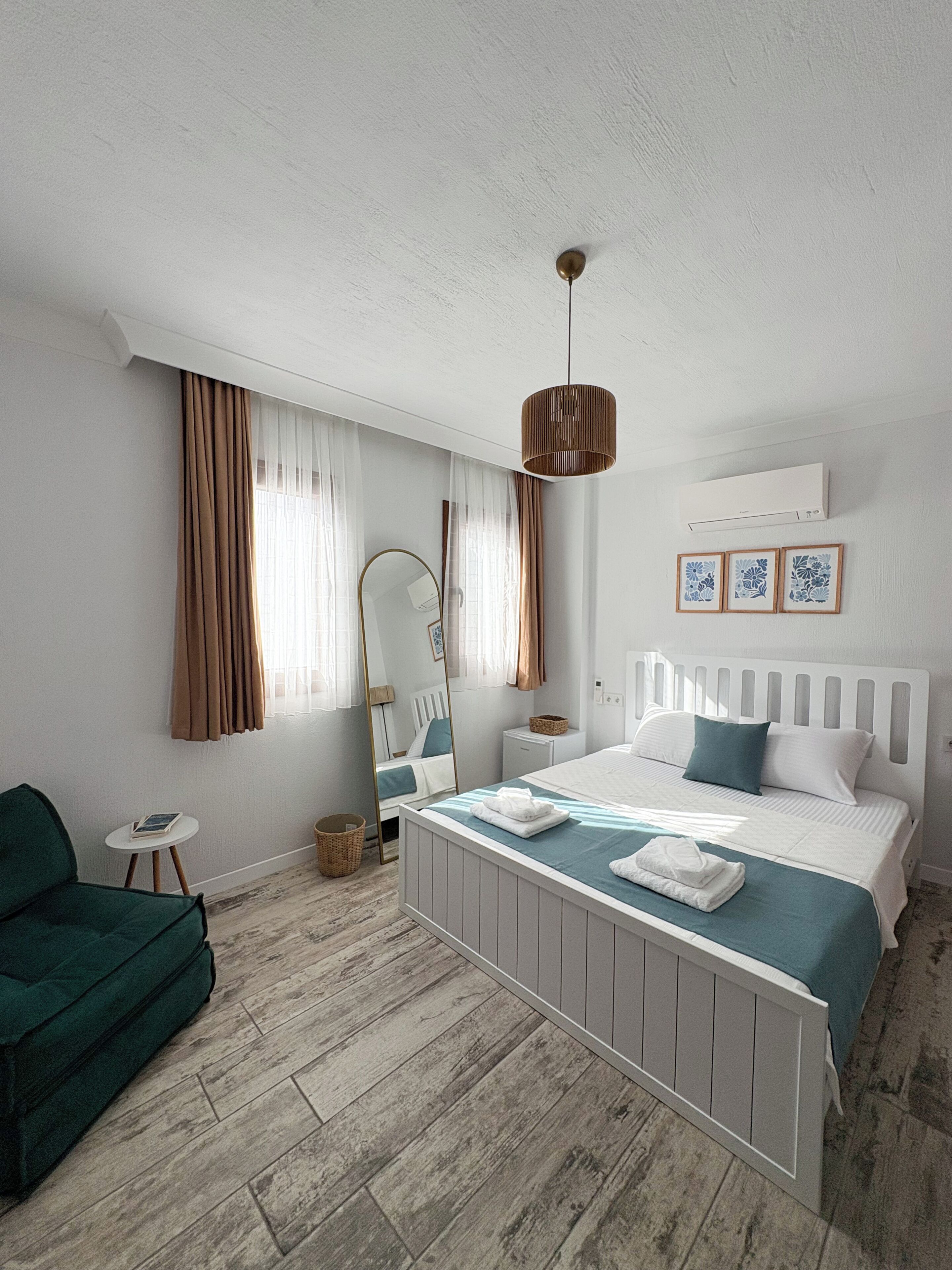 Quarto com cama, mesas de cabeceira, espelho, luminária pendente e janela com cortinas.