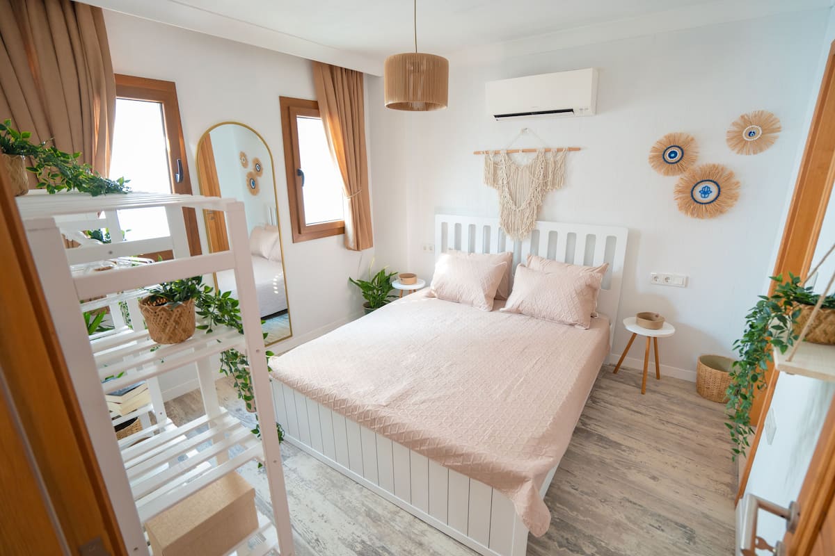 Une chambre avec un lit superposé, une table de chevet, une décoration murale en macramé et une fenêtre avec des rideaux.