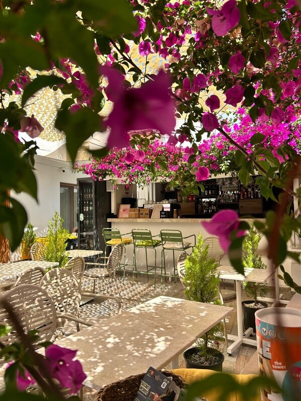 Un espacio al aire libre con mesas y sillas, una zona de bar con estantes, y vistas a un edificio con un toldo decorado.