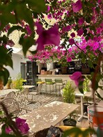Un espacio al aire libre con mesas y sillas, una zona de bar con estantes, y vistas a un edificio con un toldo decorado.