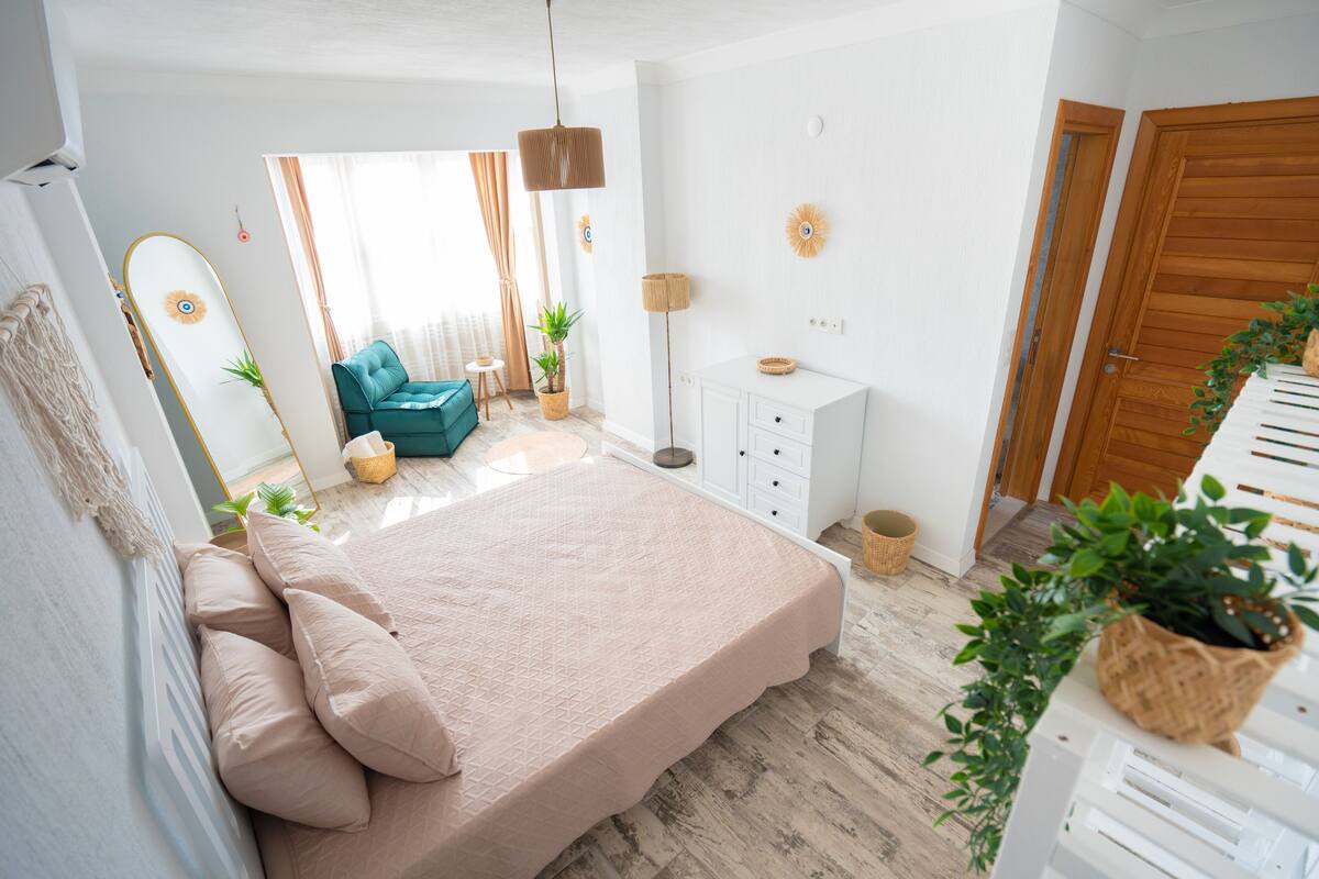 Une chambre à coucher avec un lit, un miroir, une commode, une plante et une fenêtre avec des rideaux.