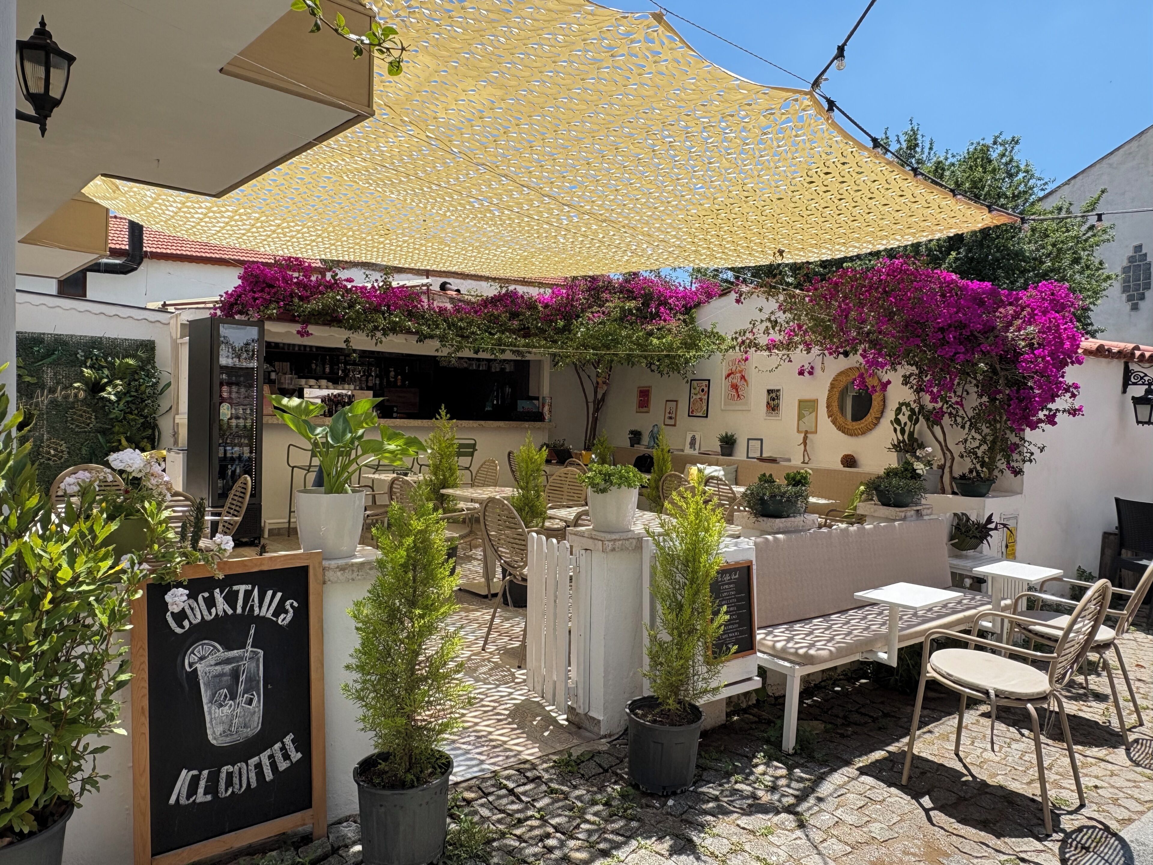 An outdoor seating area with tables and chairs, a sign for cocktails and ice coffee, and a covered section with a yellow canopy.