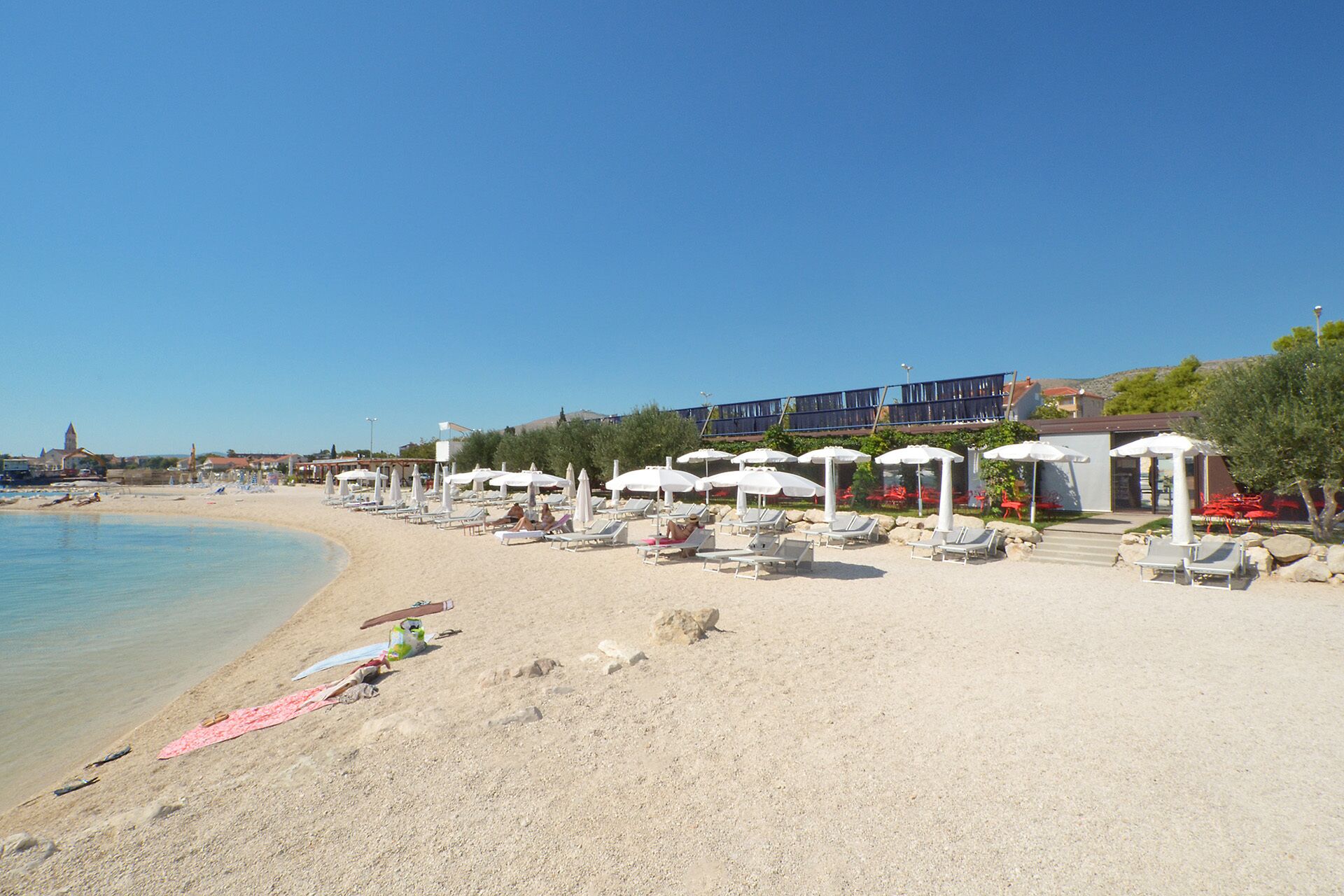 A beach with sun loungers and umbrellas, a clear sky, and buildings in the background.