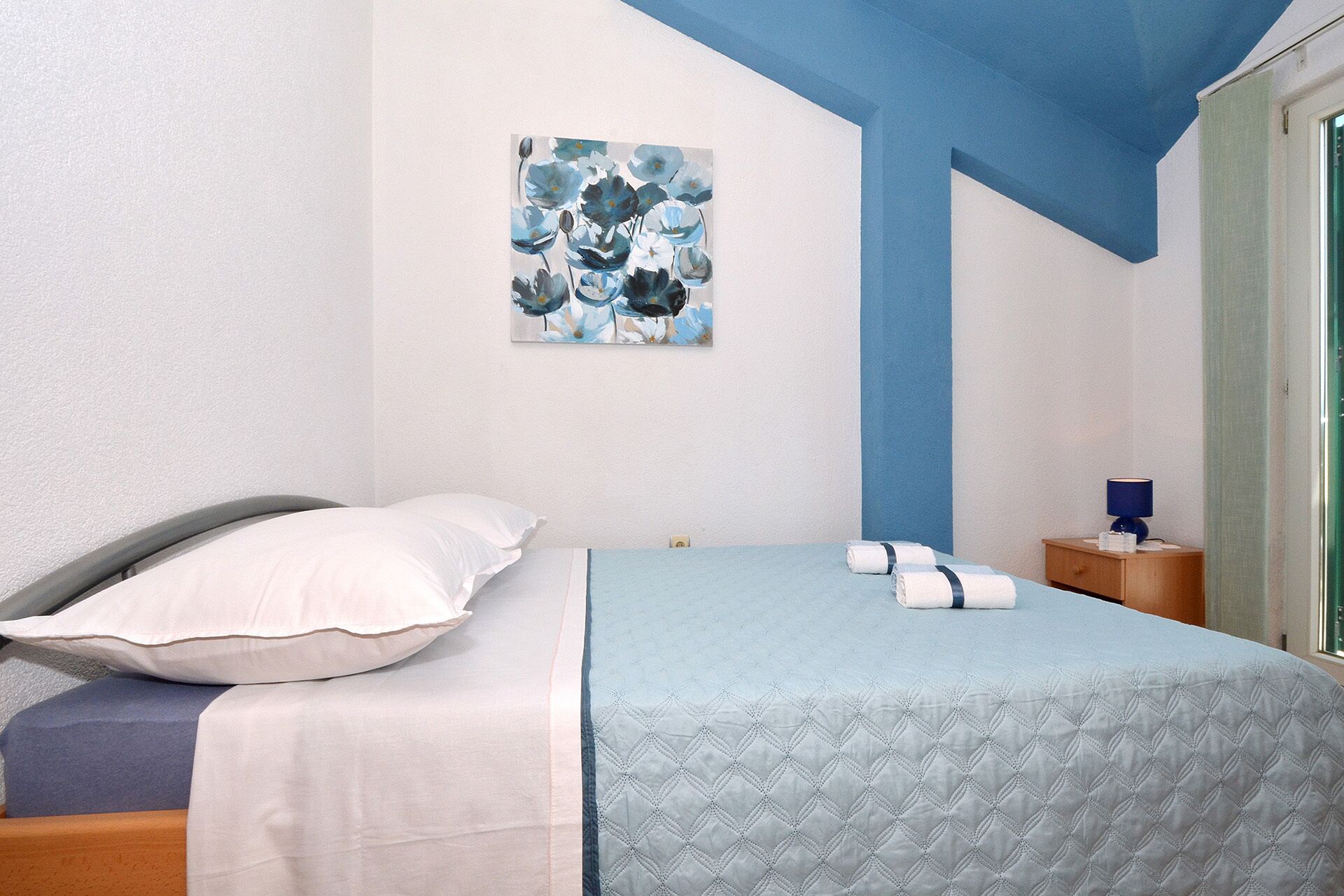 Una camera da letto con un letto, cuscini, un comodino, una lampada e un quadro appeso al muro.