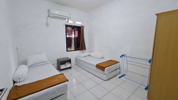 Une petite chambre propre, avec deux lits, une table de chevet, une fenêtre avec des rideaux et un radiateur.