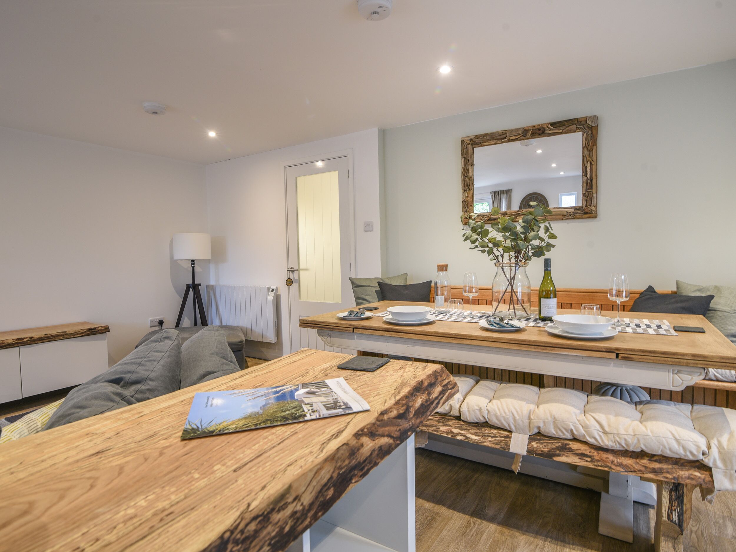 A modern, well-lit room with a wooden dining table set for two, a comfortable sofa, a lamp, and a mirror on the wall.