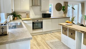 Une cuisine moderne avec des meubles blancs, un four encastré, un micro-ondes et un évier. On y trouve un îlot central en bois, sur lequel sont posés un saladier rempli de fruits et un pot de fleurs.