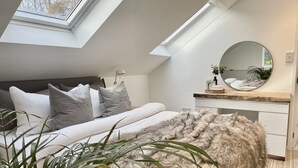 Une chambre avec une lucarne, un lit avec une literie grise et blanche, un tapis moelleux et une table de chevet avec un miroir et de petites plantes.