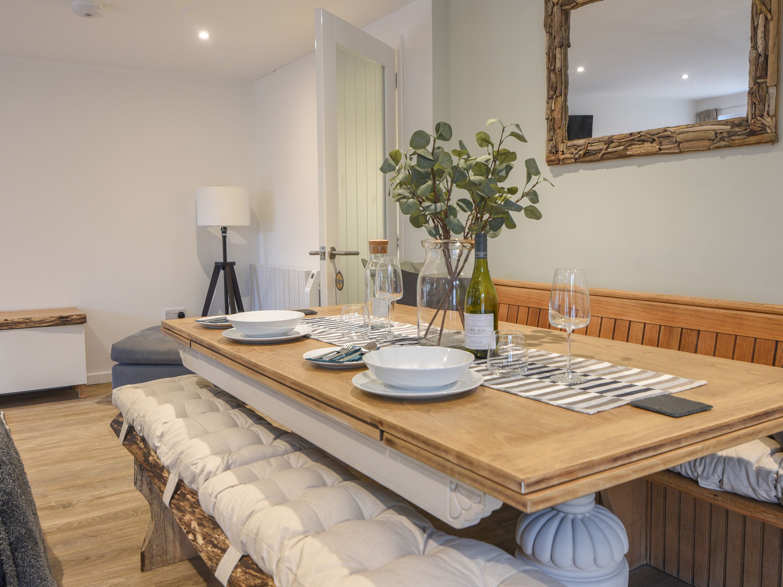 A dining area with a wooden table set for four, a bench with cushions, a mirror on the wall, and a plant on the table.