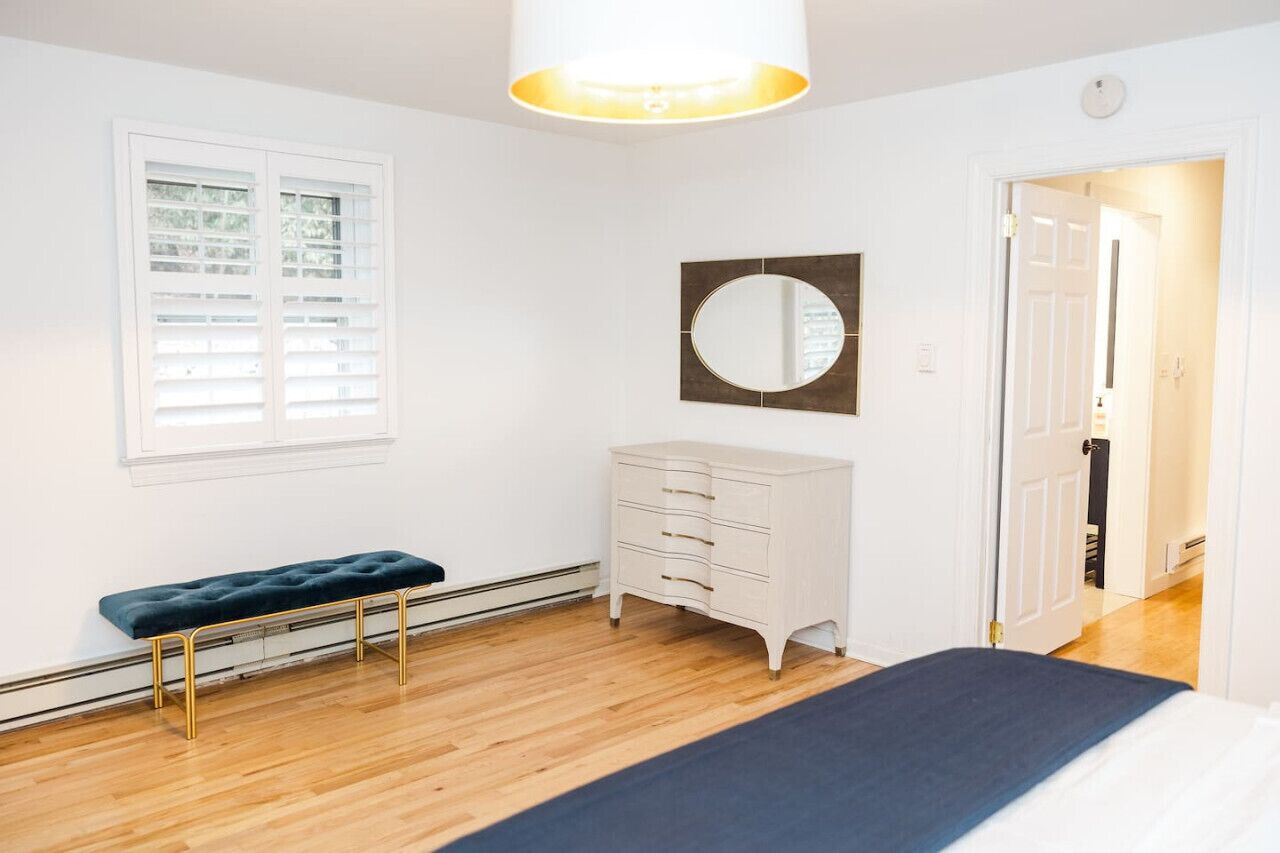 Een slaapkamer met een houten vloer, een witte linnenkast, een bankje, een ronde spiegel en een deur naar een andere kamer.