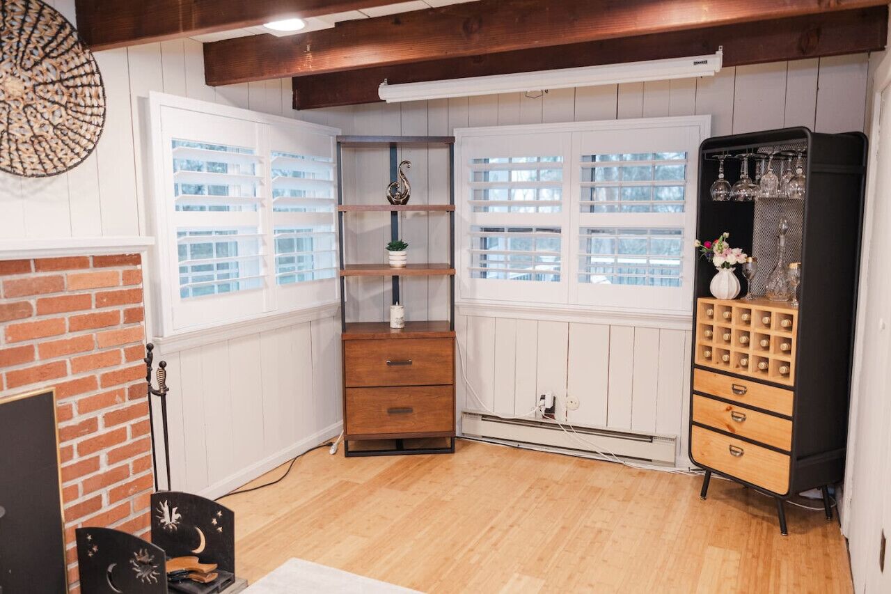 A room with a brick fireplace, white paneled walls, a wooden floor, a small wooden dresser, a black cabinet with drawers, and a window with white shutters.